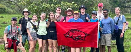 2025 Hogs Aroad picture: students holidng razorback flag in front of greenery